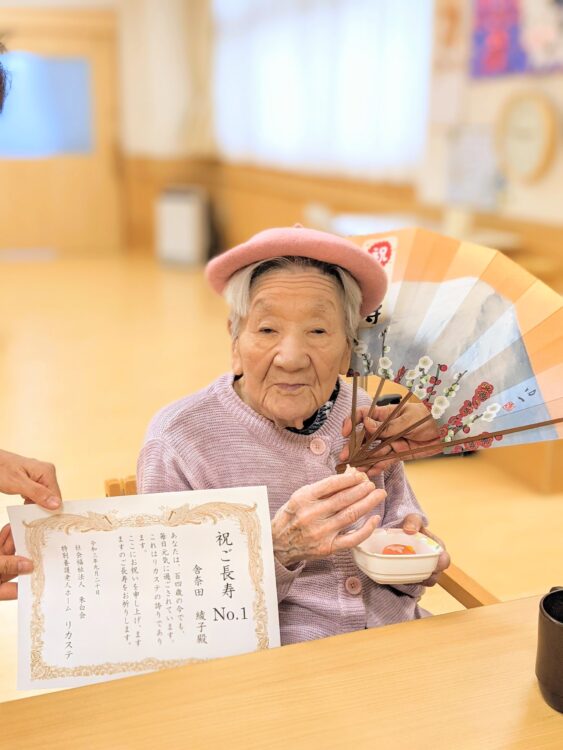 介護施設で表彰状を受け取る高齢女性入居者。ピンク帽子をかぶり笑顔で記念撮影。